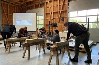 Ein Foto, das mehrere Männer beim Arbeiten an großen Holzbalken in einer geräumigen, hellen Werkstatt mit weißen Toren zeigt. Im Hintergrund ist ein Mann zu sehen, der sitzend Gitarre spielt - neben ihm ist eine Leinwand aufgebaut, auf welche ein Liedtext projiziert wird.