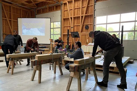 Ein Foto, das mehrere Männer beim Arbeiten an großen Holzbalken in einer geräumigen, hellen Werkstatt mit weißen Toren zeigt. Im Hintergrund ist ein Mann zu sehen, der sitzend Gitarre spielt - neben ihm ist eine Leinwand aufgebaut, auf welche ein Liedtext projiziert wird.