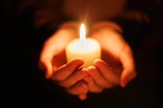 Eine von zwei Händen gehaltene, weiße Kerze, die in der Dunkelheit leuchtet.
