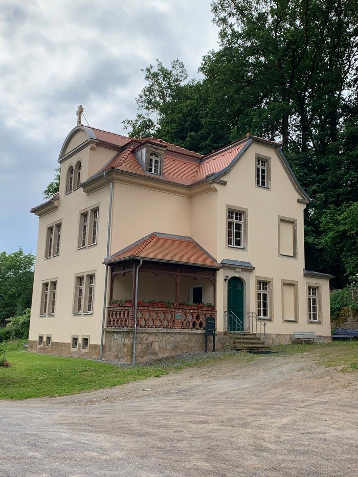 Pfarrhaus Hohenfichte mit hellgelber Fassade, blassroten Dachziegeln und Steinkreuz auf dem Giebel.
