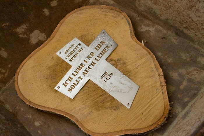 Auf einer Holzscheibe befestigtes Metallkreuz mit folgender Inschrift: "Jesus Christus spricht: 'Ich lebe und ihr sollt auch leben.' (Johannes 14,19)".